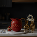 Red ceramic pitcher with strawberry pattern on a marble surface, with gold vases and strawberries in the background.